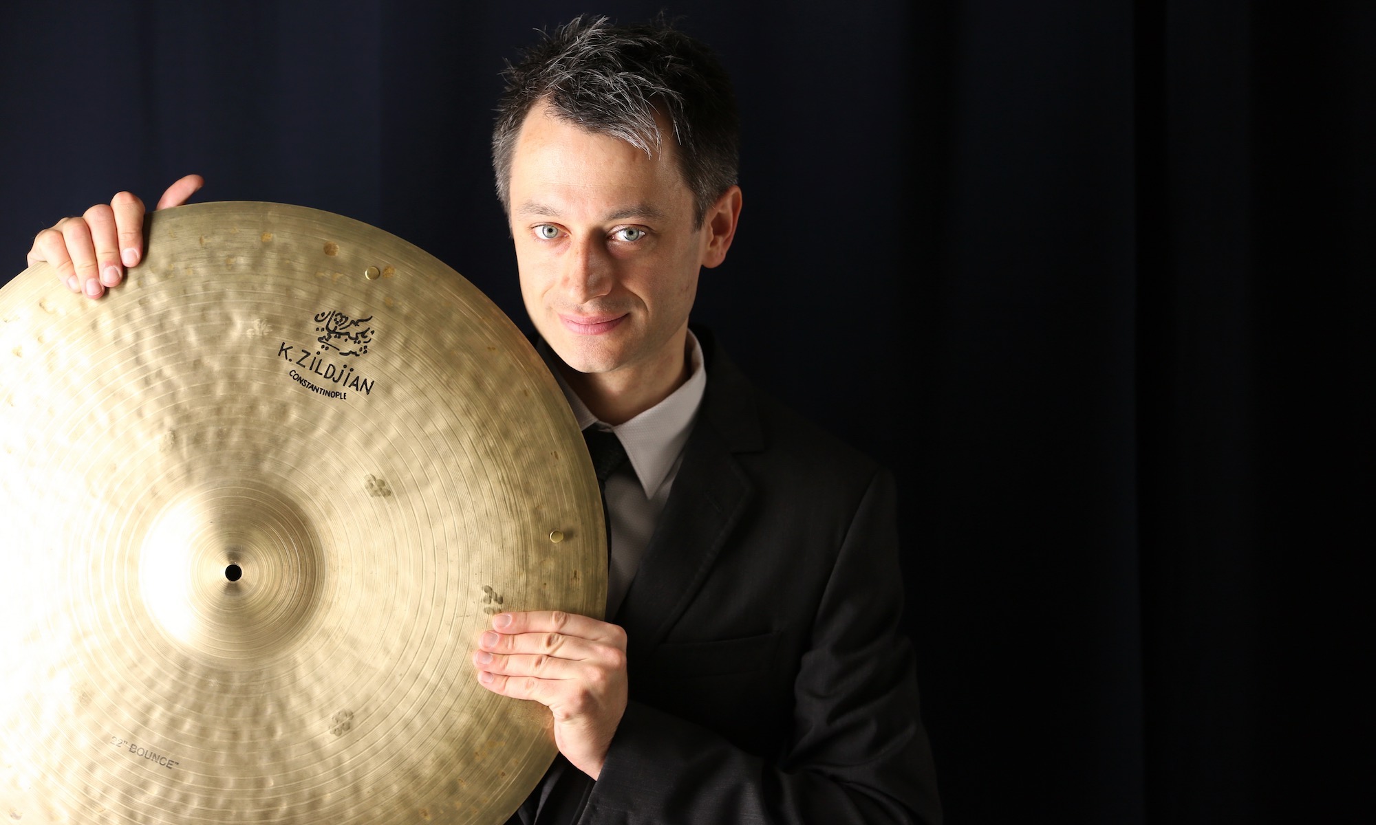 Dennis Frehse is holding a Zildjian Bounce Ride in front of a black background for his website homepage.